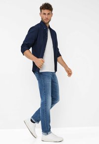 Camicia a maniche lunghe color navy sopra una t-shirt bianca, abbinata a jeans azzurri chiari e sneakers bianche, con una vestibilità comoda e maniche arrotolate.