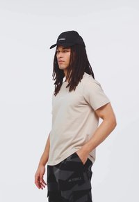 Camiseta de manga corta beige con un pequeño logo, combinada con pantalones cortos de camuflaje negros que presentan un logo, y una gorra negra con visera curva.