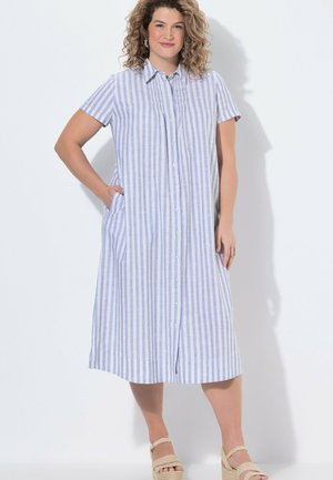 Mujer con cabello rizado, vestida con un vestido camisero de rayas verticales azules y blancas y sandalias beige de cuña, de pie con una mano en el bolsillo.