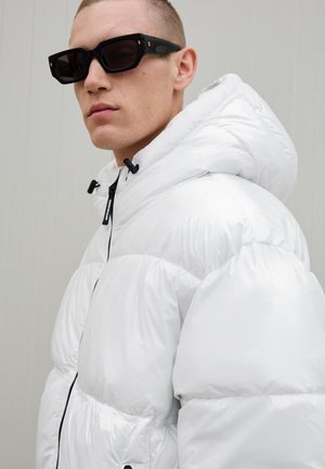 Weißer Pufferjacke mit Kapuze und Reißverschluss, ausgestattet mit elastischen Bündchen und Kordelzug. Das Modell trägt schwarze eckige Sonnenbrillen.