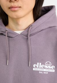 Vijolična kapuca s sprednjim logotipom, vrvicami z kovinskimi konicami in teksturirano tkanino. Blagovna znamka "ellesse" je jasno prikazana.