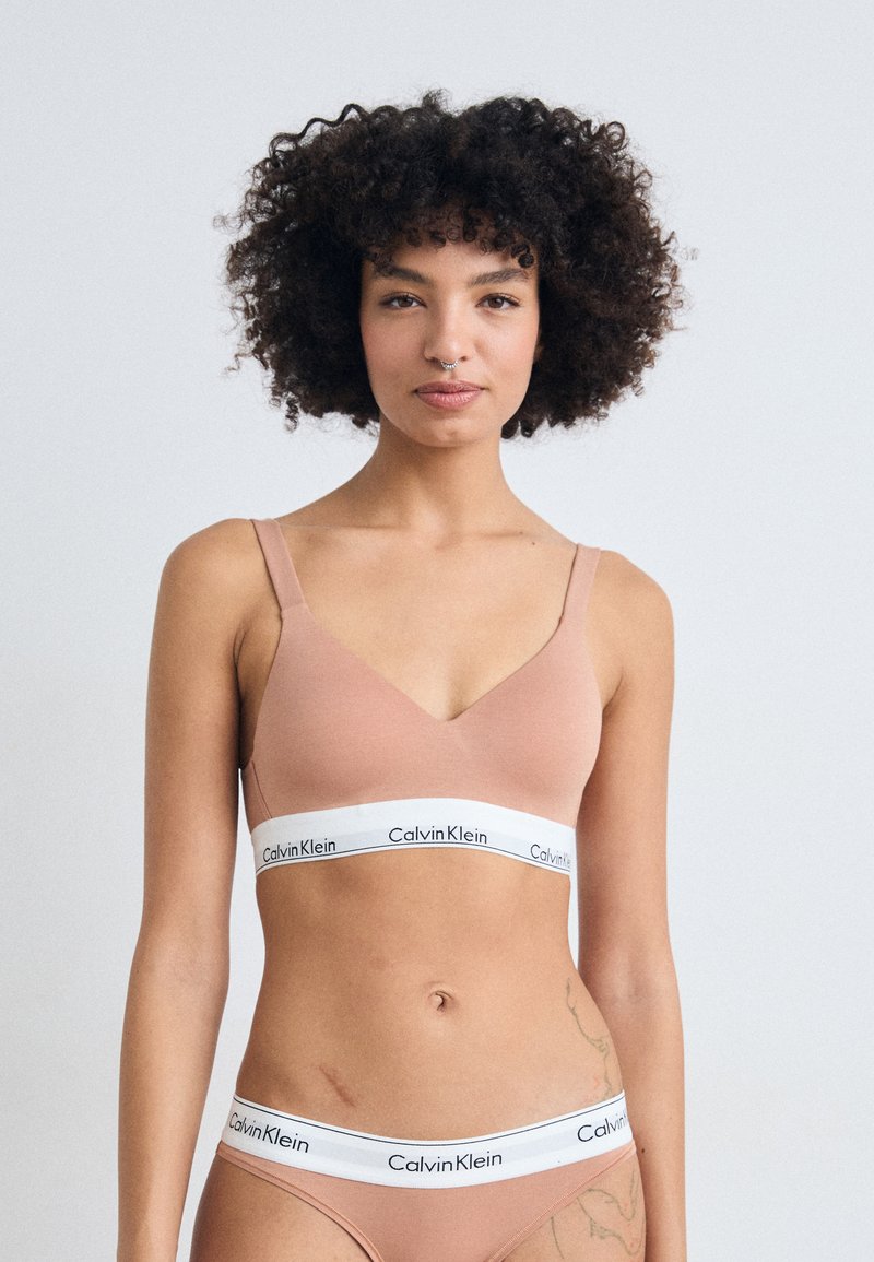 Bralette inalámbrico de Calvin Klein en suave tejido nude con un diseño en forma de V, que presenta una banda blanca con texto de logo en negro.
