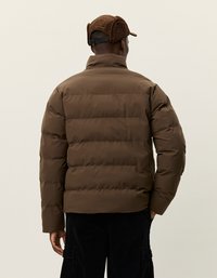 Brun pufferjakke med horisontal quilting, høy krage og tettsittende mansjetter. Brukt med en brun caps som har øreklapper.