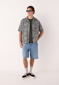 Camicia a maniche corte nera e bianca con motivo paisley sopra una t-shirt verde oliva, abbinata a pantaloncini di denim azzurro e sneakers nere.