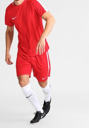Mann in rotem Nike-Fußballtrikot und weißen Nike-Socken, der auf einem einfarbigen Hintergrund läuft oder sich vorwärts bewegt.