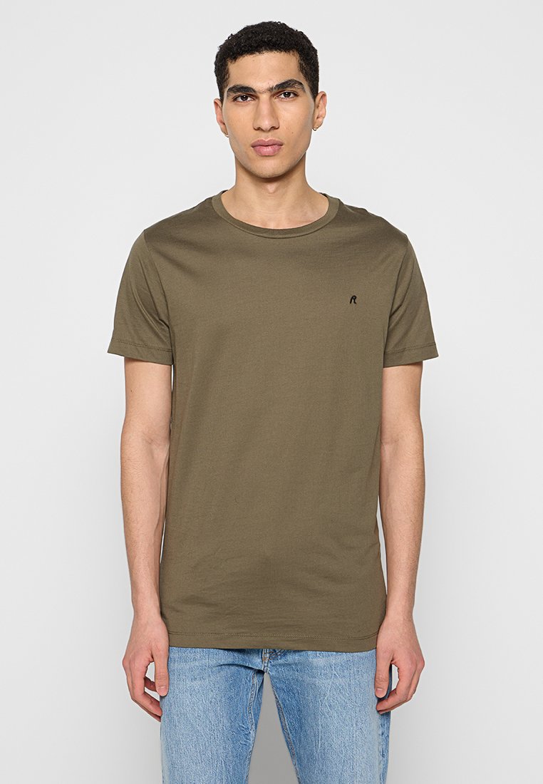 Olivegrünes Baumwoll-T-Shirt mit Rundhalsausschnitt, kurzen Ärmeln und einem kleinen schwarzen Logo auf der linken Brust. Getragen mit hellblauen Jeans.