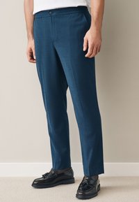 Man wearing blue tailored trousers, dark tassel loafers, and a tucked-in white shirt, standing on light carpet against beige wall.