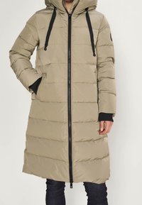Beige long padded winter coat with hood and black zipper, worn over black sleeves and dark jeans. One hand in pocket, other relaxed.