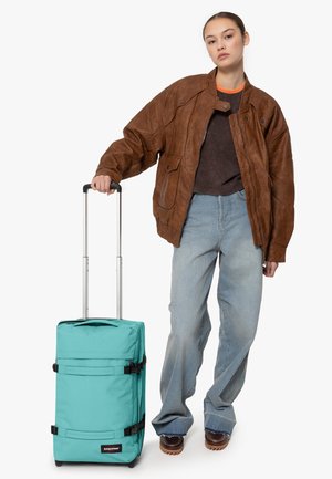 Rullande resväska i tealtyg med svarta remmar, i kombination med en brun läderjacka och lösa blå jeans. Handtaget är utdraget.