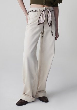 Kremowe jeansy o szerokich nogawkach z paskiem z wzorzystą chustą, zestawione z ciemnymi plecionymi butami, pokazane na stojącej osobie na jednolitym tle.