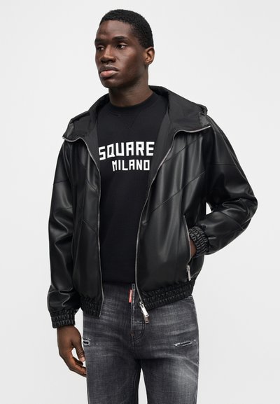 Fekete műbőr dzseki kapucnival, cipzárral és elasztikus mandzsettákkal. Egy fekete, "SQUARE MILANO" feliratos kerek nyakú pulóver fölött viselve. Szürke, kopottas hatású farmernadrág.