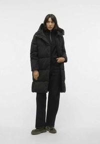 Sieviete, tērpta garā melnā pufīša mētelī ar kapuci, melnā rāvējslēdzēja džempers, tumšās platās bikses un masīvas zaļas zābaki, stāv pret baltu fona.