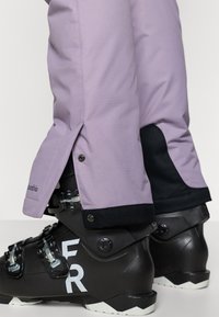 Violetas sniega bikses ar sānu atverēm, melniem akcentiem un Columbia zīmolu, kopā ar melniem slēpošanas zābakiem ar sudraba sprādzēm un baltu uzrakstu.