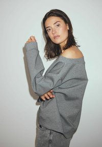 Grijze oversized sweatshirt met off-shoulder ontwerp, geribbelde manchetten en zachte textuur, gecombineerd met hoge taille spijkerbroek.