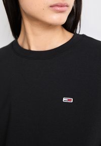 Žena v černém tričku s kulatým výstřihem, na levé hrudi má malé obdélníkové logo v červené, bílé a modré barvě, zobrazená od krku až po horní část trupu.