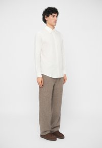 Mann står iført en langermet hvit skjorte, løse beige bukser og brune snørestøvler foran en ensfarget hvit bakgrunn.