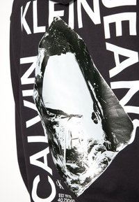 Černý oděv s bílým textem "CALVIN KLEIN JEANS" kolem velkého, lesklého, abstraktního krystalového tvaru a malým textem "cK EST 1978".