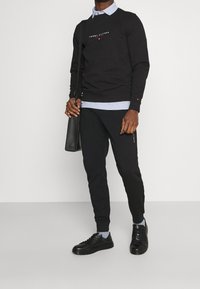 Sort sweatshirt med logo, lagdelt over en lyseblå skjorte med krave. Sorte bukser og sorte sneakers fuldender outfittet. Stilfuld lædertaske i hånden.