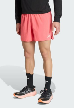 Mężczyzna noszący koralowo-różowe szorty sportowe, czarną koszulkę z długim rękawem, czarne skarpetki Adidas oraz czarne i pomarańczowe buty do biegania stoi na jednolitym tle.