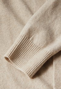 Beige gebreide trui met geribbelde manchetten en een zachte, gestructureerde afwerking, met lichte kleurvariaties door de stof heen.