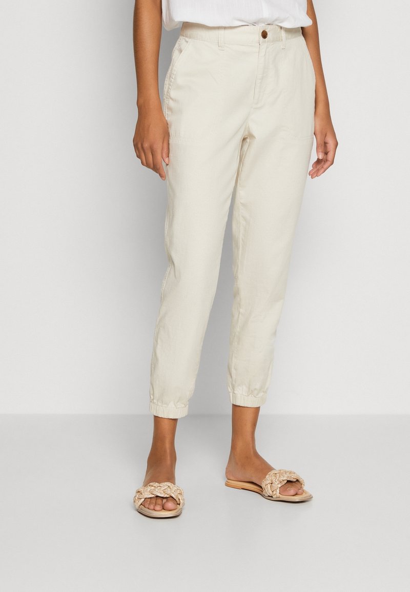 Femme portant un pantalon beige fuselé avec des poignets élastiques et des sandales tressées beiges, debout devant un fond clair uni.
