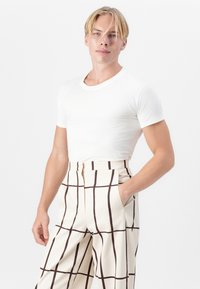 T-shirt bianca aderente abbinata a pantaloni color crema con un motivo a griglia marrone e tasche laterali. I pantaloni hanno una chiusura con zip.