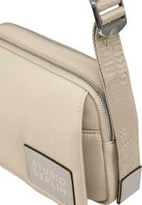 Beige crossover-veske laget av glatt stoff, med glidelås, justerbar stropp med gravert detaljer, og et logo-patch.