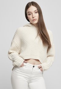 Kremfarget strikket cropped hettegenser med bred ribbestruktur og vide ermer. Kombinert med høyhalsede hvite jeans, med frontlommer og knappelukking.