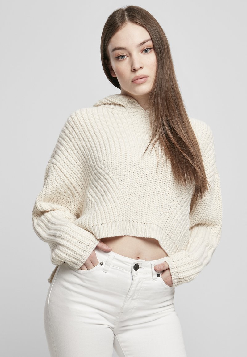 Kremfarget strikket cropped hettegenser med bred ribbestruktur og vide ermer. Kombinert med høyhalsede hvite jeans, med frontlommer og knappelukking.