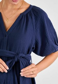 Abito midi blu navy in tessuto texturizzato, con maniche a sbuffo corte, scollo a V e dettaglio in vita con laccio. Accento con collana dorata.