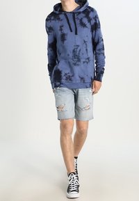Mies, kierrätetty sininen sido-tie-dye huppari, revityt vaaleat farkkushortsit ja mustat korkeavartiset tennarit kävelee tasaisen valkoista taustaa vasten.
