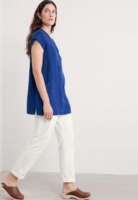 Tunika in lino blu con maniche corte, chiusura a bottoni frontale, tasche laterali, abbinata a pantaloni bianchi a sigaretta e zoccoli marroni; taglio semplice e rilassato.