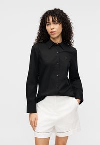 Μαύρο πουκάμισο με κουμπιά από βαμβακερό μείγμα με μυτερό γιακά, μακριά μανίκια και μικρό λογότυπο στο στήθος, σε συνδυασμό με άσπρο σορτς.