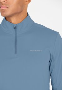 Ljusblå kvartdraget prestandatröja med en strukturerad yta. Har en hög krage och "ENDURANCE"-logotyp i silver på vänster bröst.