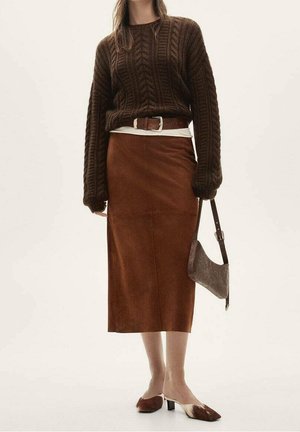 Femme portant un pull en maille torsadée brun foncé, une jupe marron taille haute, une ceinture assortie, un sac à main texturé marron et des chaussures à talons bas marron et blanc.