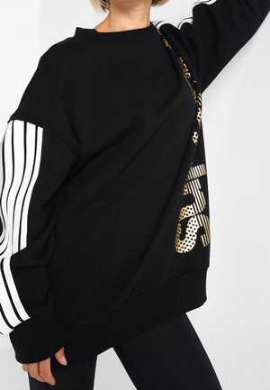 Femme portant un sweat-shirt noir oversize avec des manches blanches rayées et un texte graphique doré sur le devant, posant devant un fond blanc.