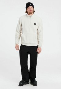 Hvid fleece-pullover med en kvart lynlås og frontlomme, parret med sorte bukser og sorte støvler, båret med en sort hue.