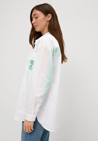 White long-sleeve shirt with green and blue stripes on the sleeves, featuring an embroidered detail on the chest and a rounded hem.