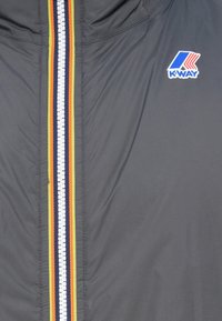 Veste K-Way en tissu gris foncé, dotée d'une fermeture éclair centrale avec des accents de rayures colorées (rouge, jaune, orange et bleu foncé).