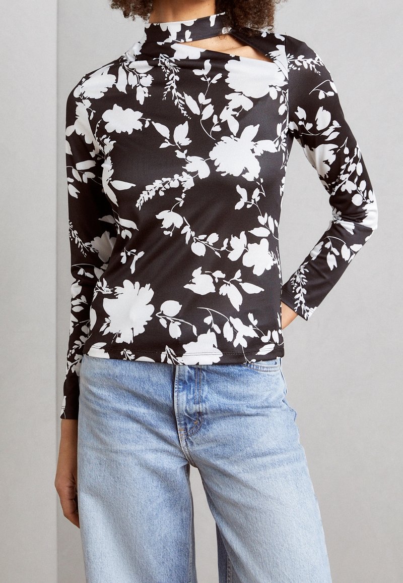 Langermige top met een zwarte stof met een witte bloemenprint, een hoge halslijn met uitsparingen en een aangesloten ontwerp. Gecombineerd met lichtblauwe jeans.