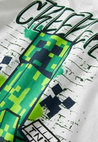 Camiseta blanca de algodón con un gráfico de personaje pixelado en verde, detalles en negro y texto superpuesto con efectos de salpicaduras.