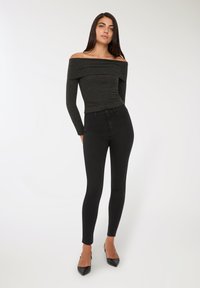 Donna con lunghi capelli scuri che indossa una maglietta a coste scura con le spalle scoperte, jeans skinny neri e tacchi a punta neri, in piedi su uno sfondo bianco.