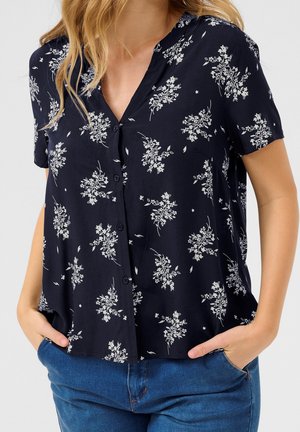 Mujer que lleva una blusa negra de manga corta con botones y patrones florales blancos metida dentro de unos vaqueros azules, manos en los bolsillos.