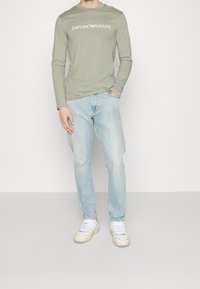 Mann trägt ein hellgrünes langärmliges Hemd von Emporio Armani, hellblaue Jeans und weiße Sneaker, steht mit einer Hand in der Tasche.