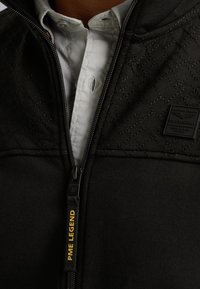 Chaqueta negra con cremallera y sección superior acolchada, que presenta un patrón texturizado y una pequeña etiqueta de la marca. El tirador de la cremallera tiene el texto "PME LEGEND".