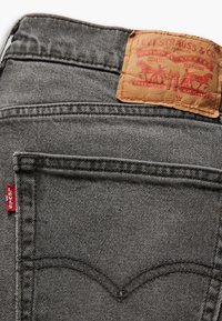 Jeans de mezclilla gris con un parche de cuero que presenta texto y gráficos en rojo. Costuras detalladas en los bolsillos traseros con una pequeña etiqueta roja.