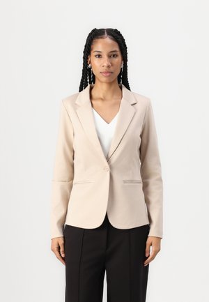 Béžový blazer s jedným gombíkom, klopovými reverzami a dvoma prednými vreckami, skombinovaný s bielym topom a čiernymi nohavicami. Hladká textúra látky.