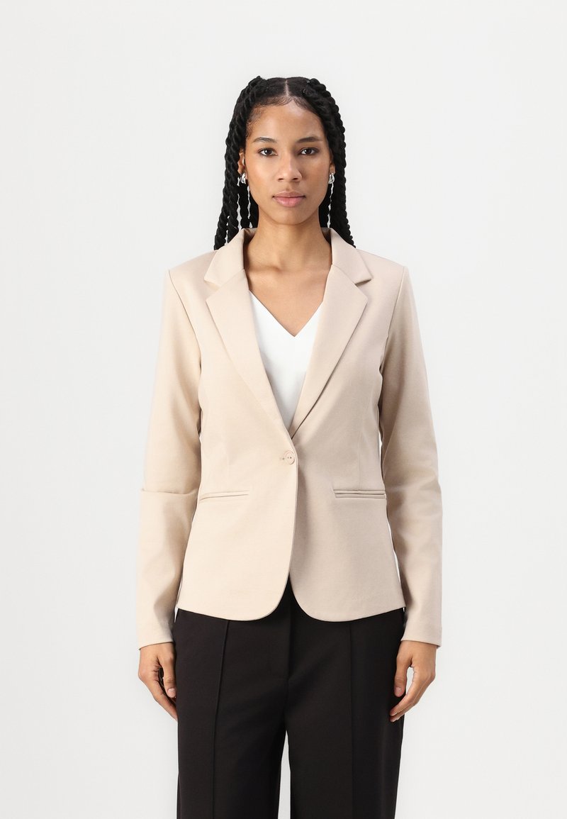 Blazer beige con un solo bottone, revers a punta e due tasche frontali, abbinato a un top bianco e pantaloni neri. Tessuto di trama liscia.