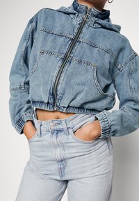 Džínová krátká bunda s kapucí, předním zipem a elastickými manžetami. Světle modrá barva, vybledlá textura a dvě velké přední kapsy.