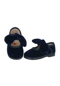 Zapatos de terciopelo azul marino con punta redonda, que presentan un gran lazo en la parte frontal y un borde elástico negro alrededor de la abertura, apoyados en una suela plana.